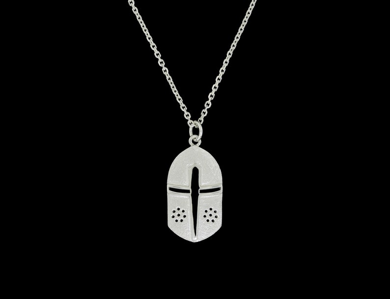 May include: A silver necklace with a pendant shaped like a knight's helmet. The helmet has a cross design and small holes around the face.