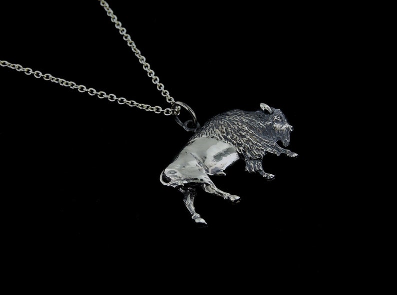 May include: A silver pendant necklace featuring a detailed bison sculpture. The bison is depicted in a running pose with its head turned to the side. The pendant is attached to a silver chain.