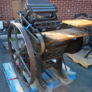 May include: A vintage printing press with a large wooden wheel and a metal frame. The press is on a wooden pallet and has a wooden platform on top.