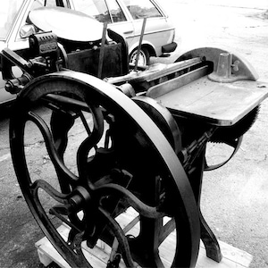 May include: Black and white image of an antique printing press. The press features a large, ornate flywheel, a flat printing surface, and various gears and levers. The machine is made of cast iron and sits on a wooden pallet.