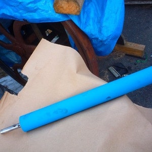 May include: A blue roller with a metal rod extending from one end. The roller is lying on a brown paper surface.