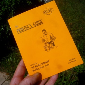 May include: A yellow booklet titled "The Printer's Guide" published by The Kelsey Company in Meriden, Connecticut. The cover features a black and white illustration of a man operating a printing press. The text "9th Edition Price 50 cents" is printed in black at the bottom right corner.