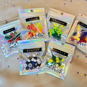 May include: Six iridescent pouches, each containing colourful beads and letter beads spelling names. The pouches are labelled with names such as "RORY", "ARLO", "ARTHUR", "GRAYSON", "BERTIE", and "ALFIE". Confetti is scattered around the pouches.