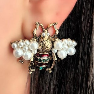 Pode incluir: Brincos de abelha dourados com detalhes de pérolas. Os brincos apresentam um design detalhado de abelha com enfeites vermelhos, verdes e pérolas. Os brincos estão presos a uma orelha.