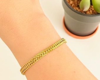 Op de afbeelding: Een goudkleurige armband met een gevlochten kettingontwerp wordt om een pols gedragen. De armband is gemaakt van glanzend metaal en heeft een textuur. Een kleine vetplant in een pot staat op de achtergrond.