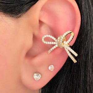 Può includere: Un orecchino a cerchio dorato a forma di fiocco, incrostato di piccoli cristalli trasparenti. Sono visibili anche due piccoli orecchini a bottone. Il gioiello è indossato sull'orecchio.