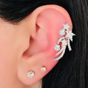 Puede incluir: Un ear cuff plateado con diseño de estrella y floral, adornado con cristales transparentes. También se ven dos pequeños pendientes de botón. La joya se encuentra sobre cabello oscuro.