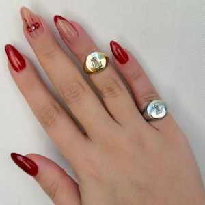 May include: A hand displaying two signet rings, one gold and one silver, each with a rectangular clear stone. The nails are painted red with a nude base and red tips. The nail polish is a deep red.