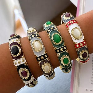 May include: Four hinged bangle bracelets featuring gold-tone metal accents, enamel detailing, and central oval cabochon stones. The bracelets display varied color combinations including black, white, green, red, and burgundy with floral motifs.