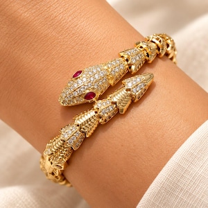 Può includere: Un bracciale dorato a forma di serpente con un corpo testurizzato e segmentato incastonato con numerose piccole pietre chiare. La testa del serpente è dettagliata con due pietre rosse ovali per gli occhi e una superficie pavé di pietre chiare.