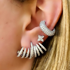 Può includere: Set di orecchini argentati con tre diversi modelli. Un ear cuff curvo, un piccolo punto luce quadrato e un design che avvolge l'orecchio. Tutti gli orecchini sono impreziositi da piccole pietre scintillanti.
