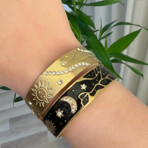 Può includere: Tre braccialetti in smalto oro e nero con disegni celesti. Il braccialetto superiore presenta un disegno di cielo stellato con strass trasparenti. Il braccialetto centrale ha un disegno del sole. Il braccialetto inferiore ha un disegno di luna e stelle.