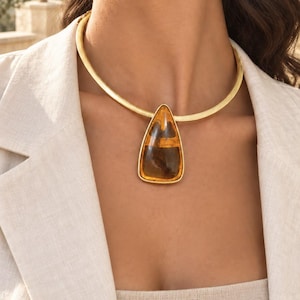 Puede incluir: Un collar tipo gargantilla de metal dorado con un gran colgante en forma de lágrima de color ámbar con inclusiones internas visibles. El collar se lleva alrededor del cuello, complementado con un pequeño pendiente de botón dorado en el lóbulo.