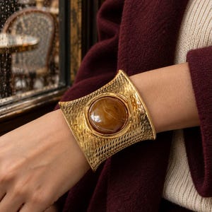 Puede incluir: Un brazalete ancho de tono dorado con una superficie metálica texturizada de patrón entrecruzado y una piedra grande, lisa y circular de color marrón engastada en el centro. El brazalete tiene una forma cóncava curva que se adapta a la muñeca.