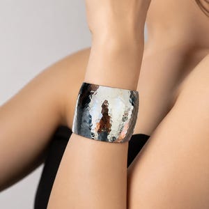 Puede incluir: Un brazalete ancho de tono plateado con una superficie martillada y texturizada que crea una apariencia reflectante y con hoyuelos. La banda de metal tiene una estructura rígida y curva que rodea la muñeca, mostrando un acabado metálico pulido.