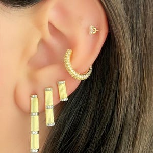 K&ouml;nnte beinhalten: Nahaufnahme eines Ohrs mit goldenen Ohrringen. Das Ohr hat einen kleinen Stecker, einen gebogenen Reifen und zwei vertikale Ohrringe. Die Ohrringe sind goldfarben mit kleinen, funkelnden Akzenten, was einen glamour&ouml;sen Look erzeugt.