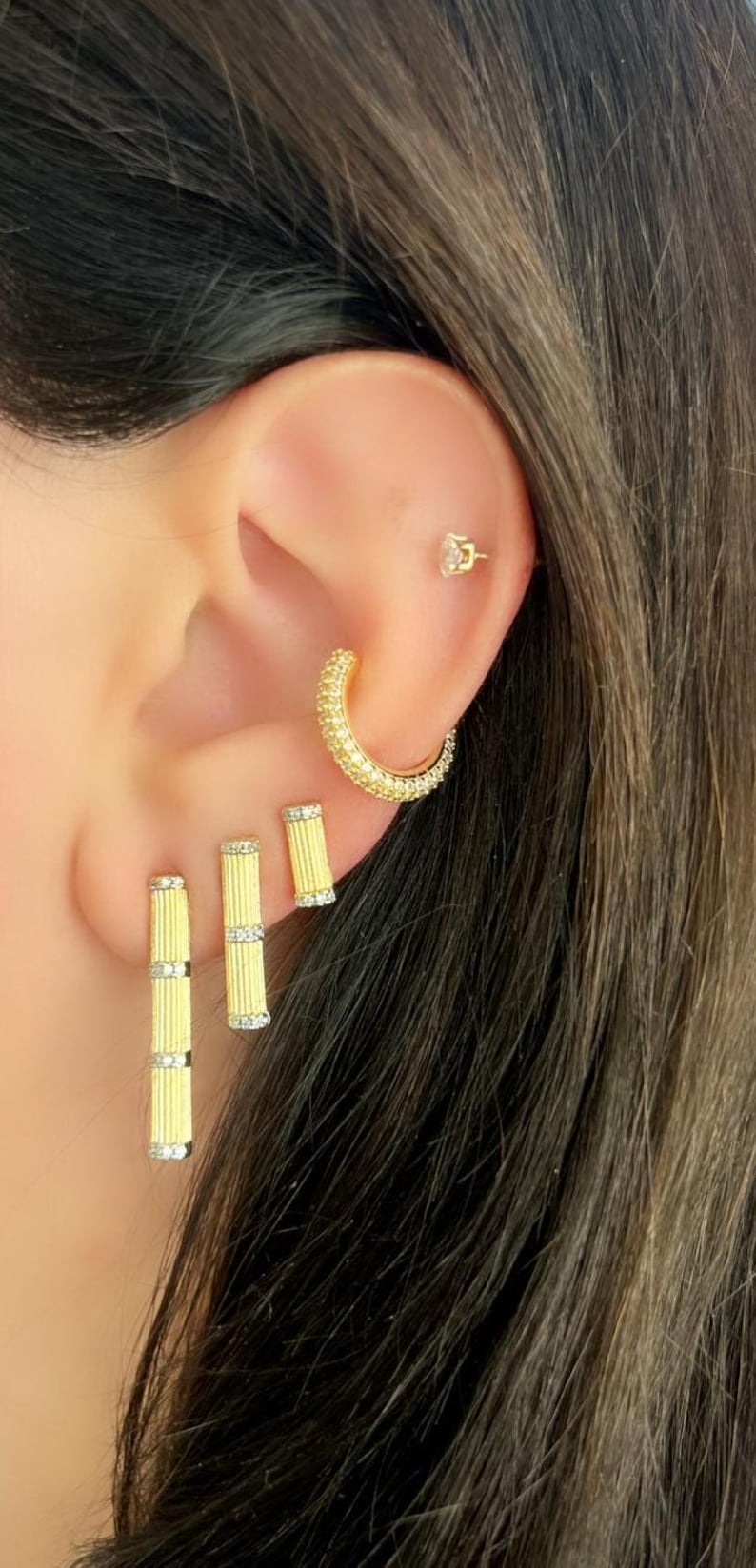 K&ouml;nnte beinhalten: Nahaufnahme eines Ohrs mit goldenen Ohrringen. Das Ohr hat einen kleinen Stecker, einen gebogenen Reifen und zwei vertikale Ohrringe. Die Ohrringe sind goldfarben mit kleinen, funkelnden Akzenten, was einen glamour&ouml;sen Look erzeugt.