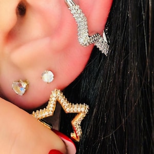 Puede incluir: Juego de pendientes con un pendiente en forma de estrella con metal dorado y cristales transparentes, un pendiente curvo plateado con cristales transparentes y dos pendientes pequeños. Los pendientes se muestran en una oreja con cabello negro.