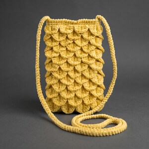 May include: A handmade, yellow crochet crossbody bag. The bag features a textured, scale-like pattern and a long, braided strap. The bag is made of a soft, yellow yarn and is suitable for carrying small items. The background is a solid gray.
