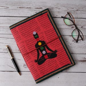 Puede incluir: Un diario rojo con un diseño de chakra, que presenta una figura meditando con símbolos de chakra coloridos. El diario tiene un borde verde y dorado, un cierre de cordón rojo y está acompañado de un bolígrafo y gafas.