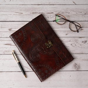 Puede incluir: Un diario de cuero marrón oscuro con diseños de nudos celtas en relieve y un cierre de metal. Un bolígrafo plateado y negro y un par de gafas con montura marrón descansan junto al diario sobre una superficie de madera clara. El diario está cerrado.