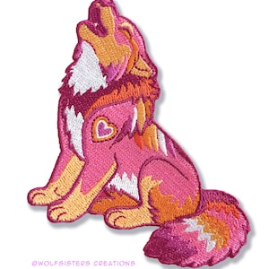 Peut inclure: Écusson brodé représentant un loup hurlant dans des tons de rose, orange et blanc. Le loup a un détail en forme de cœur. Le texte "@wolfsisters creations" est en bas.