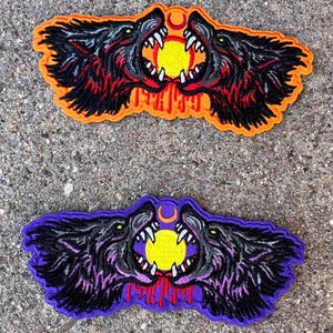 Puede incluir: Dos cabezas de lobo negras con los colmillos descubiertos, enfrentadas, con un sol amarillo y una luna creciente entre ellas. Las cabezas de lobo están bordadas en un parche con fondo naranja.