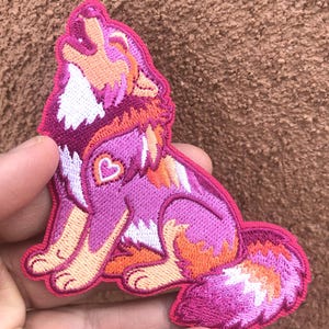 May include: Embroidered patch featuring a howling wolf in shades of pink, orange, and white. The wolf has a heart-shaped design on its chest. The patch is likely for clothing or accessories.