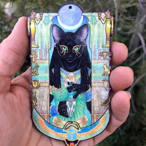 Puede incluir: Un adorno de madera con un gato negro de ojos verdes, que lleva un collar de oro y un vestido verde. El gato sostiene un pequeño jarrón blanco con una flor dentro. El adorno está decorado con símbolos egipcios y una luna creciente.