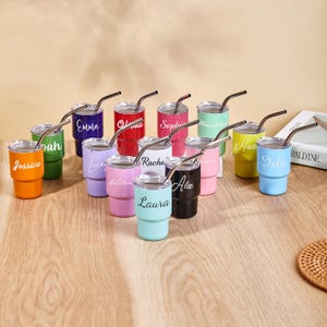 May include: A collection of colorful, personalized stainless steel tumblers with silver straws. Each tumbler is a different color, including orange, green, purple, pink, and blue, and features a name in white script. The tumblers are arranged on a wooden surface.