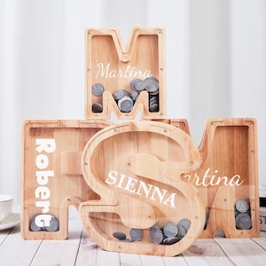 May include: Wooden letter-shaped coin banks, each filled with coins. The letters are M, S, and M, with names like "Martina", "Sienna", and "Robert" printed on them. The banks are made of light-colored wood.