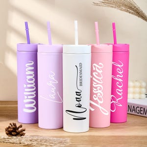 May include: Five colourful, personalised tumblers with straws. The tumblers are in shades of lavender, light purple, white, pink, and magenta. Each has a name in a contrasting script font. The white tumbler is labelled "Bridesmaid".