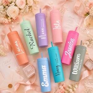 Op de afbeelding: Een verzameling gekleurde bekers met rietjes, gepersonaliseerd met namen in witte letters. De pastelkleuren variëren, met namen als "Rachel" en "Isla". De bekers zijn gerangschikt met bruiloftdecoraties.