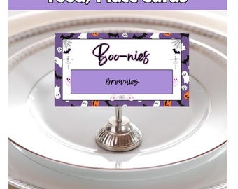 Op de afbeelding: Een Halloween-thema tafelkaart met de tekst "Boo-nies" en "Brownies" op een witte en paarse achtergrond, versierd met vleermuizen, geesten en spinnenwebben. De kaart wordt vastgehouden door een zilveren metalen standaard, geplaatst op een wit bord.