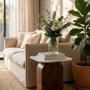 Könnte beinhalten: Ein Wohnzimmer mit beigem Sofa und Kissen. Ein Beistelltisch aus Holz mit Marmorplatte trägt eine Vase mit weißen Rosen und zwei Büchern. Rechts steht eine Topfpflanze. Auf dem Boden liegt ein Jute-Teppich.