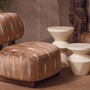 Puede incluir: Una escena de salón moderno con una silla estampada marrón y crema con estructura de madera y dos mesas auxiliares color crema. La silla tiene respaldo redondeado y asiento estampado. Las mesas auxiliares tienen un diseño geométrico.