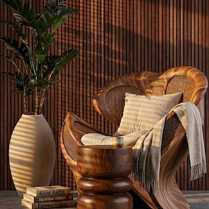 Op de afbeelding: Een houten stoel met een geweven rugleuning en een gestreept kussen, een kleine houten bijzettafel en een grote vaas met een plant. Boeken met de titels "Alvar Aalto", "Japanese Rattan" en "Eames" zijn naast de vaas gestapeld. De achtergrond is een houten muur.