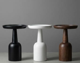 Op de afbeelding: Drie moderne bijzettafels in zwart, wit en bruin. Elke tafel heeft een rond blad en een cilindrische voet. De zwarte tafel is geverfd, de witte tafel is glad en de bruine tafel is van hout.