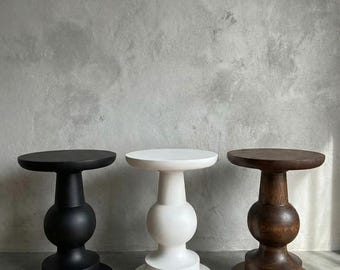 Op de afbeelding: Drie decoratieve bijzettafels. Eén is zwart, één is wit en de derde is donkerbruin hout. Elke tafel heeft een rond blad en een sculpturale voet. De tafels staan op een grijze ondergrond. Waarschijnlijk van hout.