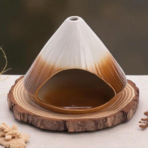 May include: A ceramic essential oil diffuser with a cone-shaped design, featuring a white top that transitions to a brown base. It sits on a natural wood slice base, creating a rustic and earthy aesthetic. The diffuser has an opening for releasing the scent.