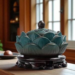 Puede incluir: Un quemador de incienso de cerámica color turquesa con forma de flor de loto, con base y tapa marrón oscuro. El quemador tiene un diseño detallado y está colocado sobre un soporte de madera. El fondo incluye una ventana y una superficie de madera.