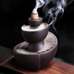 Puede incluir: Un quemador de incienso de cerámica marrón oscuro con un diseño escalonado. Un cono de incienso encendido se encuentra en la parte superior, emitiendo humo blanco que cae en cascada por los niveles. El quemador descansa sobre una superficie de madera sobre un fondo negro.
