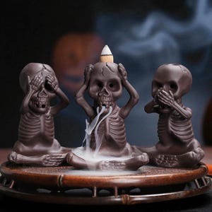 Puede incluir: Tres figuras de calaveras de cerámica marrón en posición sentada, cada una con un gesto diferente: tapándose los ojos, los oídos y la boca. La calavera central sostiene un cono de incienso, emitiendo humo blanco. Sobre una bandeja de madera.