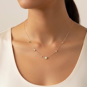 May include: Delicate gold necklace featuring a series of gemstones. The necklace includes a central opal, surrounded by smaller diamonds, emeralds, and other gemstones. The necklace is worn around the neck.
