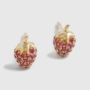 May include: A pair of gold-colored strawberry stud earrings. Each earring features a gold-colored stem and a body encrusted with small, round, pink gemstones. The earrings are set against a white background.