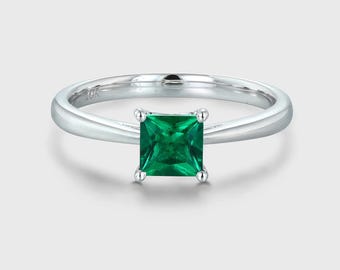 May include: A silver ring with a square, emerald-cut green gemstone. The band is smooth and polished, with a subtle inscription near the top. The gemstone is held in place by four small prongs. The ring is set against a light background.