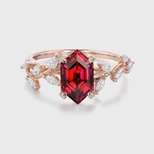 Op de afbeelding: Roségouden ring met een grote, zeshoekige, gefacetteerde rode edelsteen als middelpunt. De ring is versierd met kleine, witte, marquise-geslepen stenen en bladvormige details op de band, wat een elegant en door de natuur geïnspireerd ontwerp creëert.