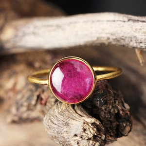 May include: A gold ring featuring a large, round, magenta-colored gemstone. The ring's band is thin and gold, complementing the vibrant gemstone. The ring is set against a natural, textured wooden background.