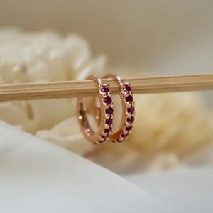 Könnte beinhalten: Roségoldene Creolen-Ohrringe mit einer Reihe kleiner, tiefroter Edelsteine. Die Ohrringe werden auf einem hellbraunen Stock vor einem weichen, verschwommenen Hintergrund präsentiert. Ein klassisches Schmuckstück.