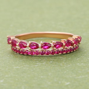 Könnte beinhalten: Ein roségoldener Ring mit zwei Reihen leuchtend rosa Edelsteinen. Die obere Reihe hat tropfenförmige Steine, während die untere Reihe mit kleineren, runden Edelsteinen besetzt ist. Der Ring ist vor einem sanften grünen Hintergrund platziert.